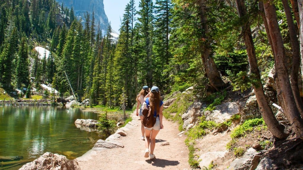 Two women walk a shaded trail beside a reflective mountain lake surrounded by tall evergreens.