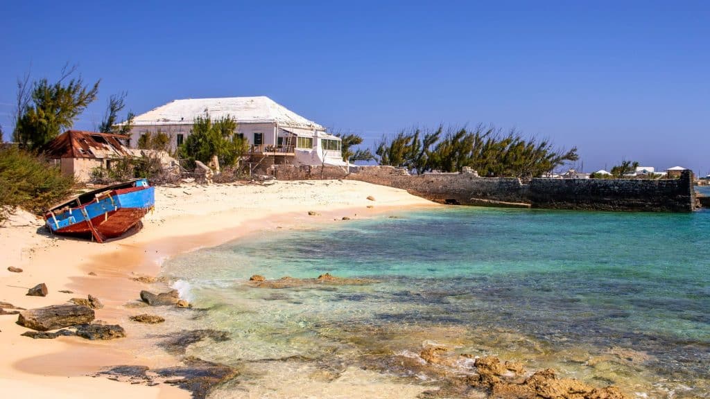 A weathered blue and red boat rests on the sandy shore beside turquoise water, with an old house and stone wall in the background.