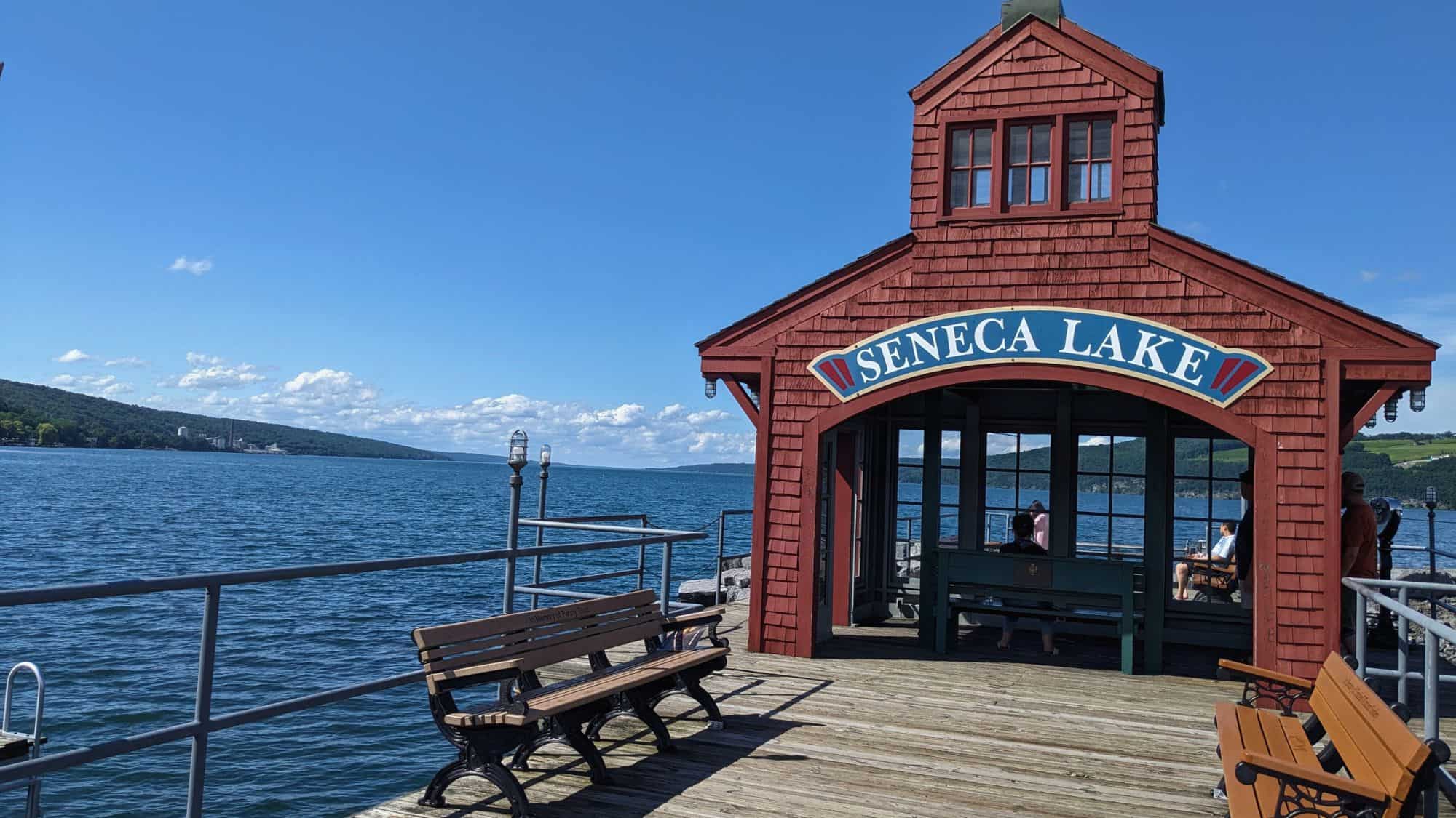 A red wooden pavilion labeled “Seneca Lake” sits at the end of a pier, overlooking calm blue waters and rolling hills in the distance.