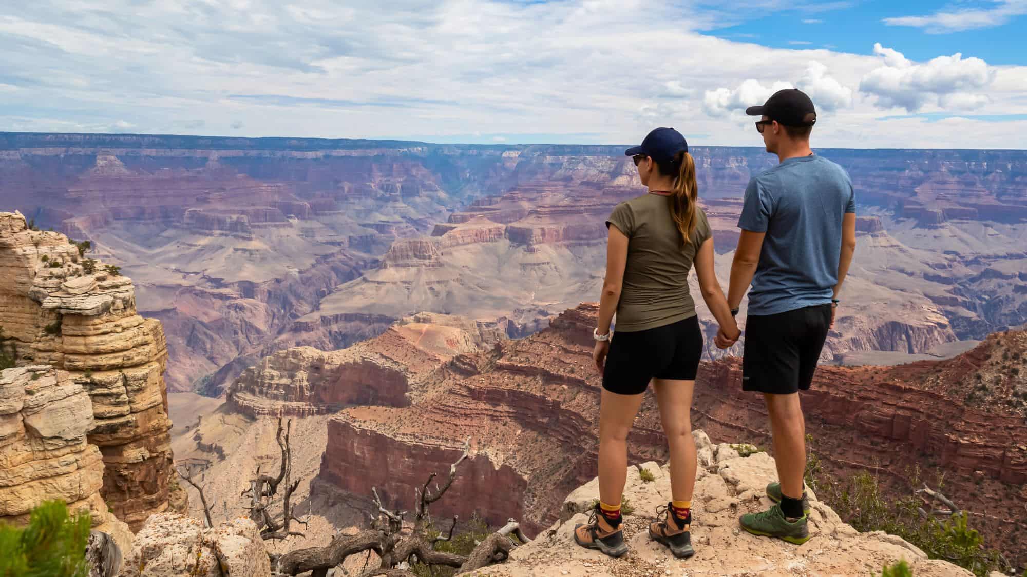 A couple stands hand in hand on a rocky edge, gazing over the vast, layered expanse of the Grand Canyon.