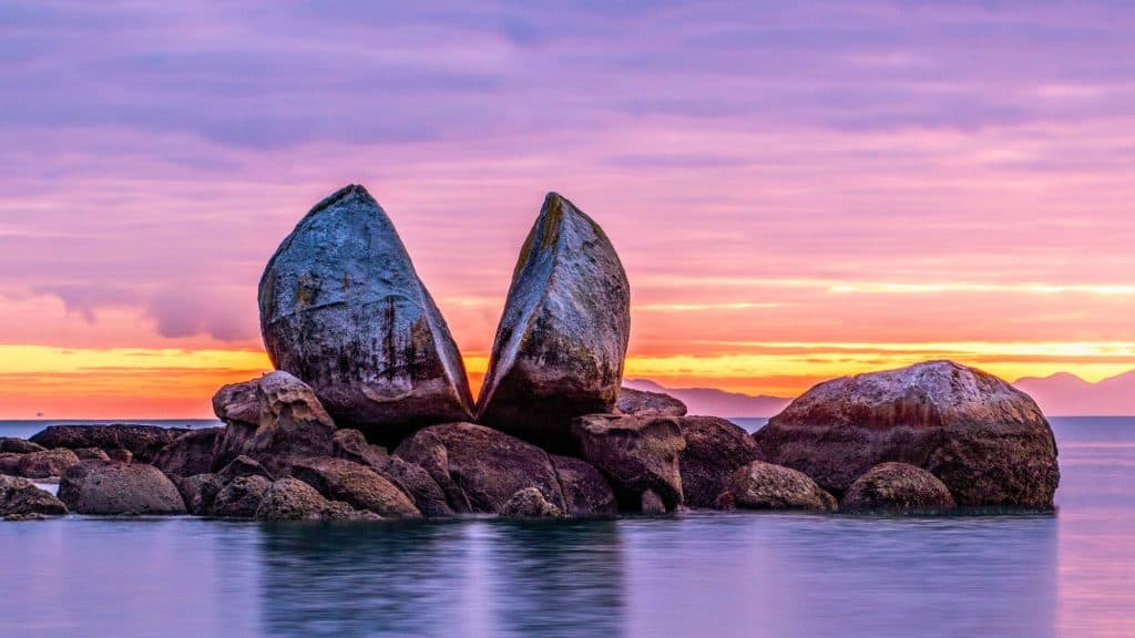 A large, naturally split boulder sits in tranquil water beneath a colorful pink-and-purple sunset sky.