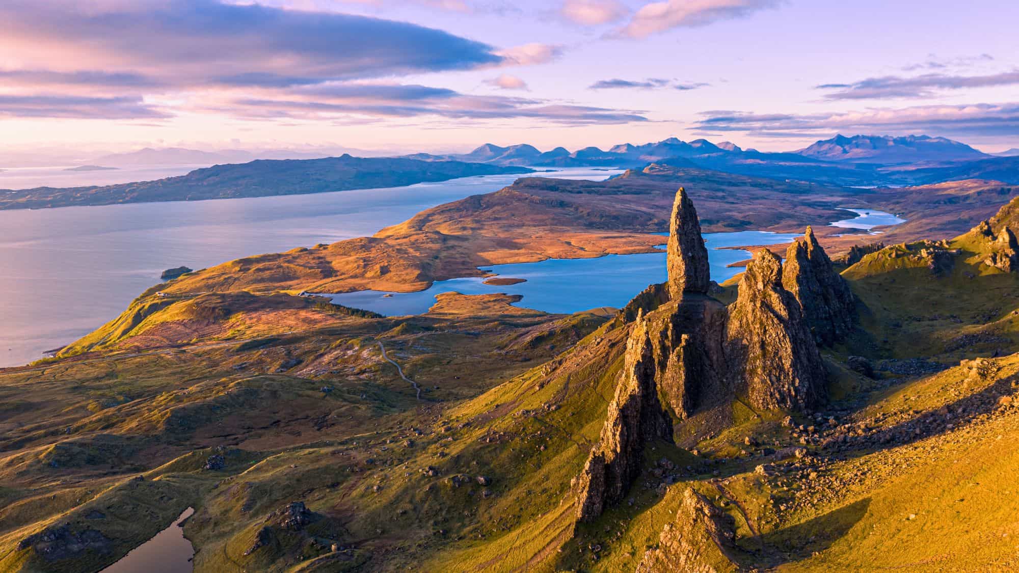 Jagged rock pinnacles rise from rolling green highlands, overlooking shimmering lochs and distant mountains in the golden light of sunrise.