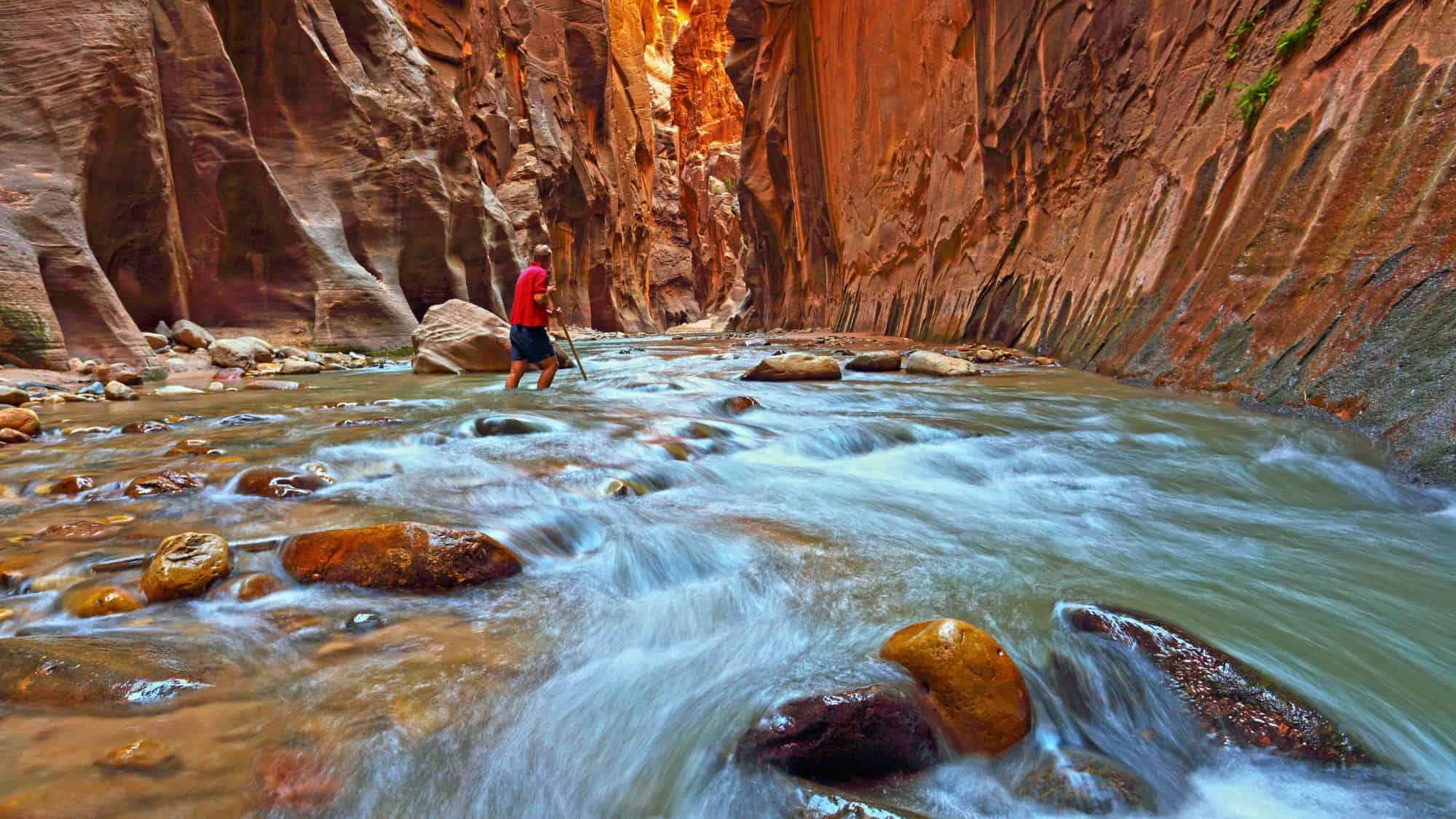 A hiker wades through the rushing waters of a narrow canyon, surrounded by towering red rock walls.