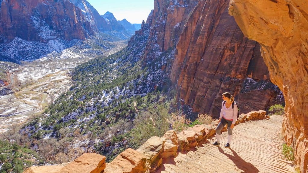 A hiker walks along a winding path carved into red rock cliffs, overlooking a snowy valley and rugged canyon landscape.