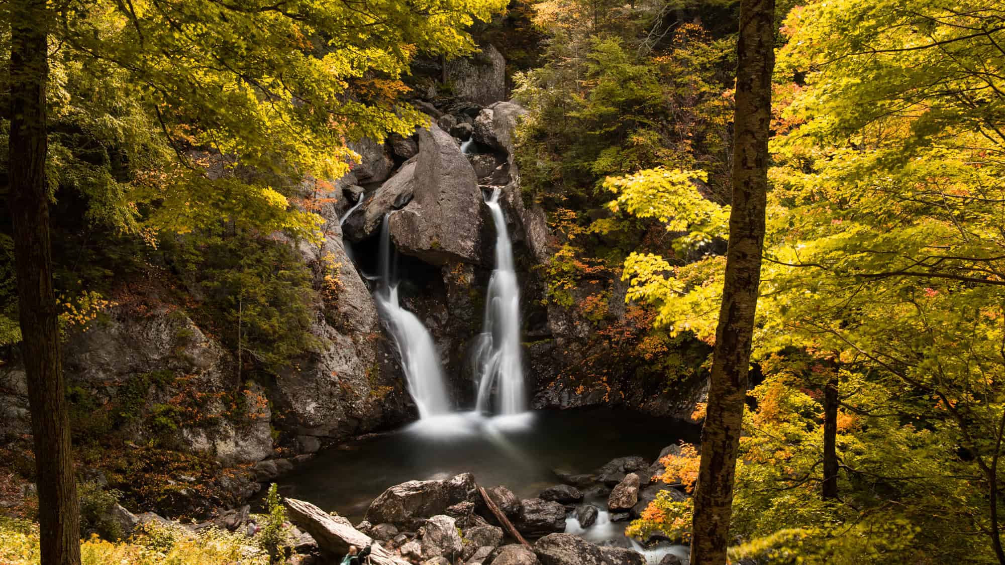 A dramatic double waterfall flows between jagged rocks into a round pool, surrounded by golden fall leaves and mossy cliffs.
