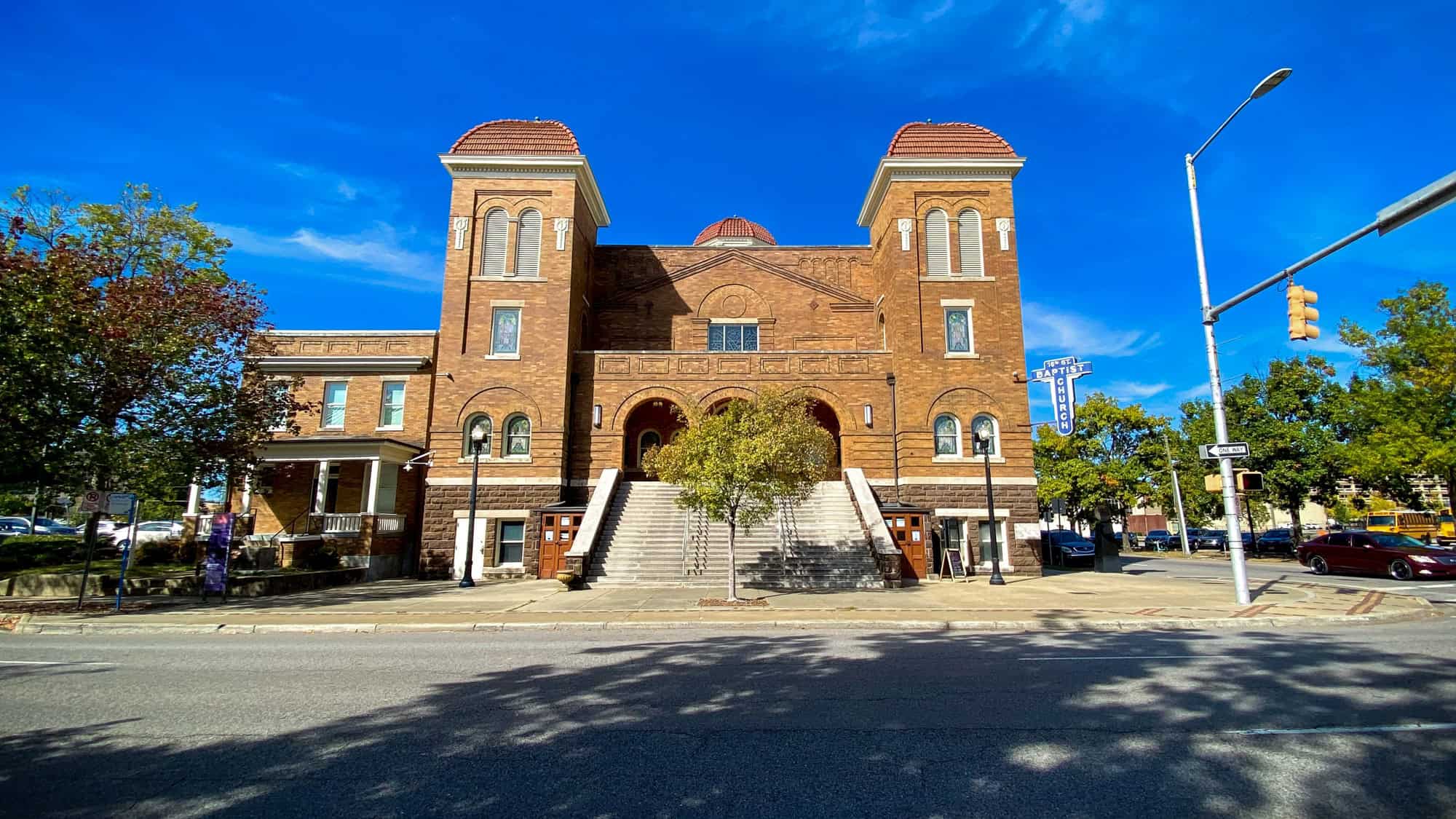 A red-brick church with twin towers and wide steps stands under a clear blue sky, marked by a sign reading “Baptist Church.”