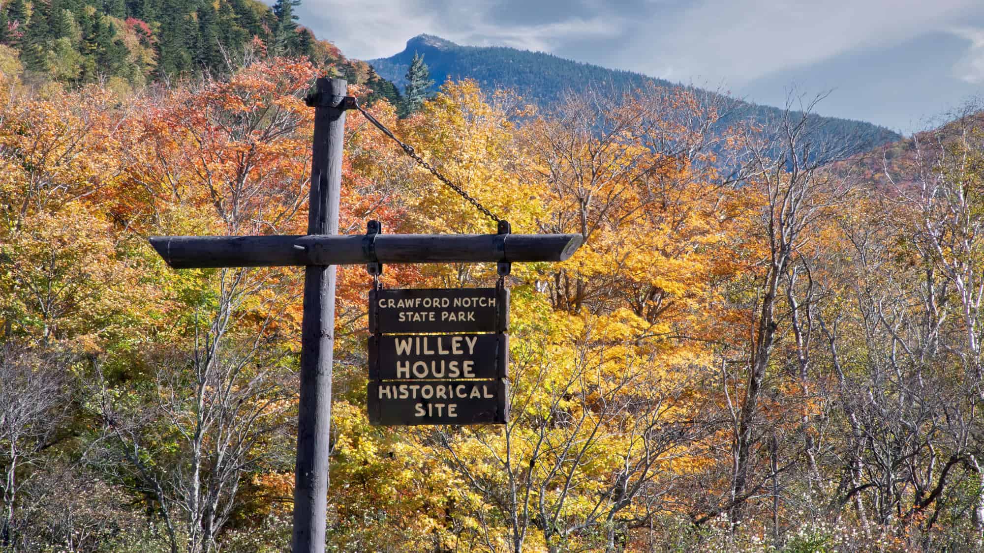 A wooden sign for Crawford Notch State Park’s Willey House Historical Site stands against a backdrop of colorful autumn foliage and mountain peaks.