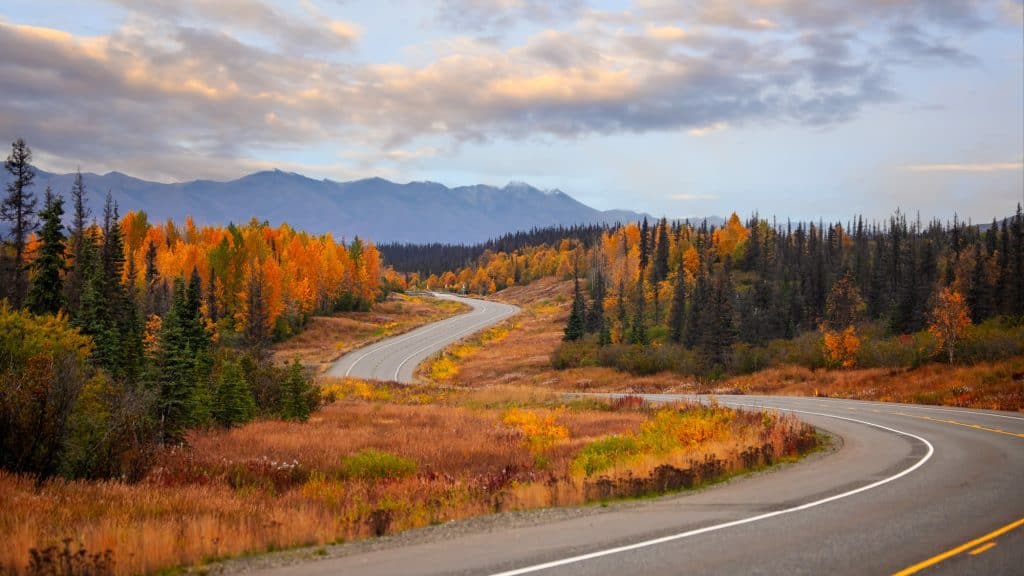 A winding highway cuts through vibrant fall foliage and evergreen forests with distant mountains under a colorful evening sky.