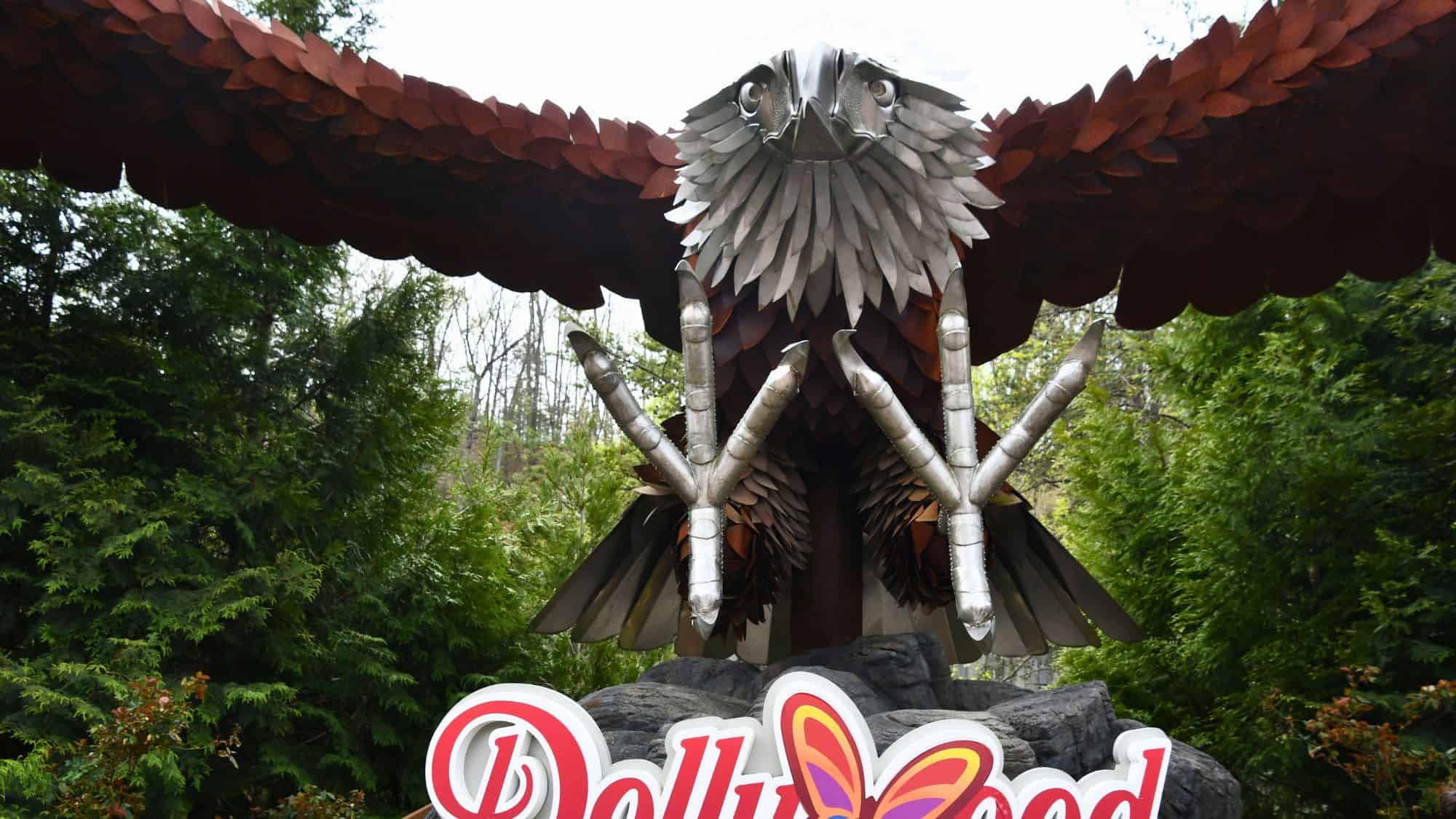 A massive metal eagle sculpture with outstretched wings towers above a Dollywood sign, set against a backdrop of lush green trees.