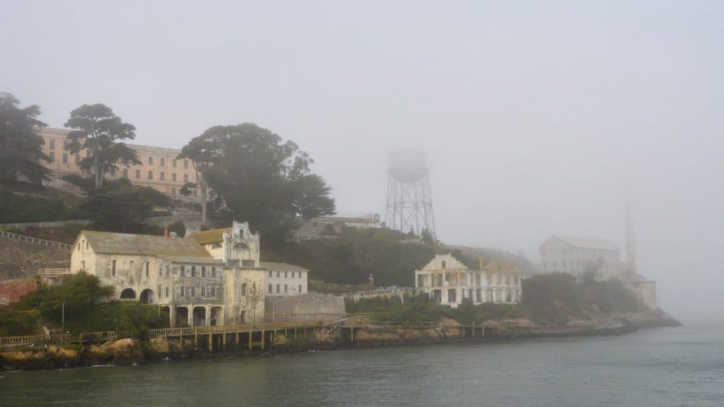 Alcatraz Island appears shrouded in thick fog, with its decaying waterfront buildings and water tower partially obscured against the gray sky.