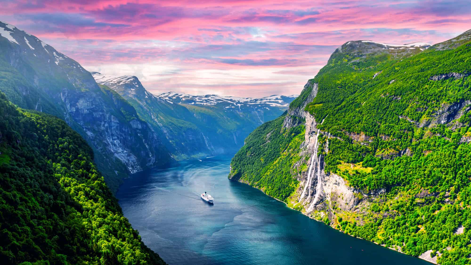 A cruise ship sails through the vivid blue waters of Geirangerfjord, flanked by lush cliffs and a tall waterfall.