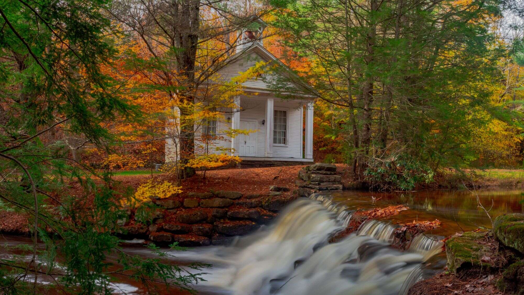 A small white chapel with a steeple sits tucked in the woods beside a gentle stream and stone dam, framed by vibrant autumn trees.