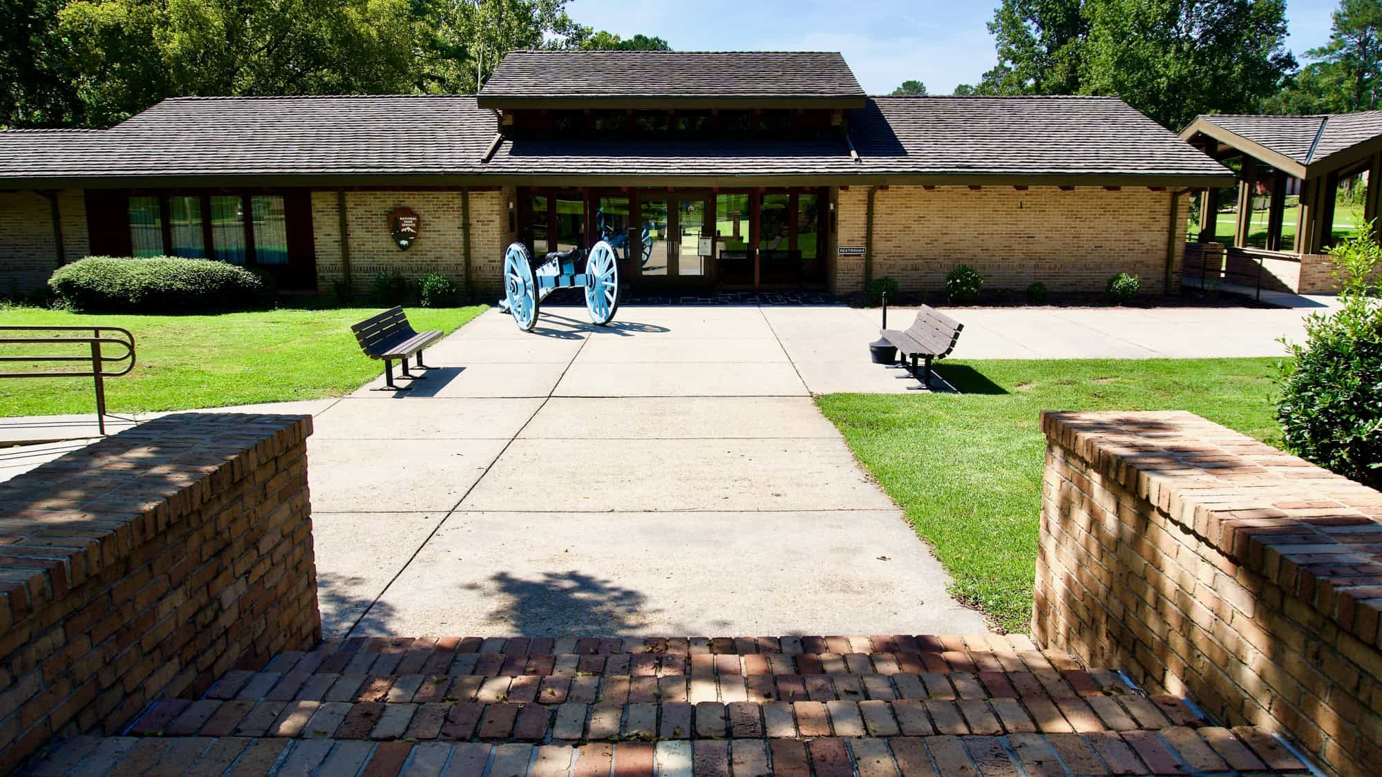 A brick pathway leads to a visitor center building with a light blue cannon displayed at the entrance.