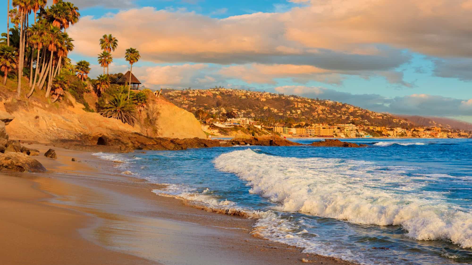 Palm trees and rugged cliffs frame turquoise waves as they crash onto the sandy shore with a coastal town in the background.