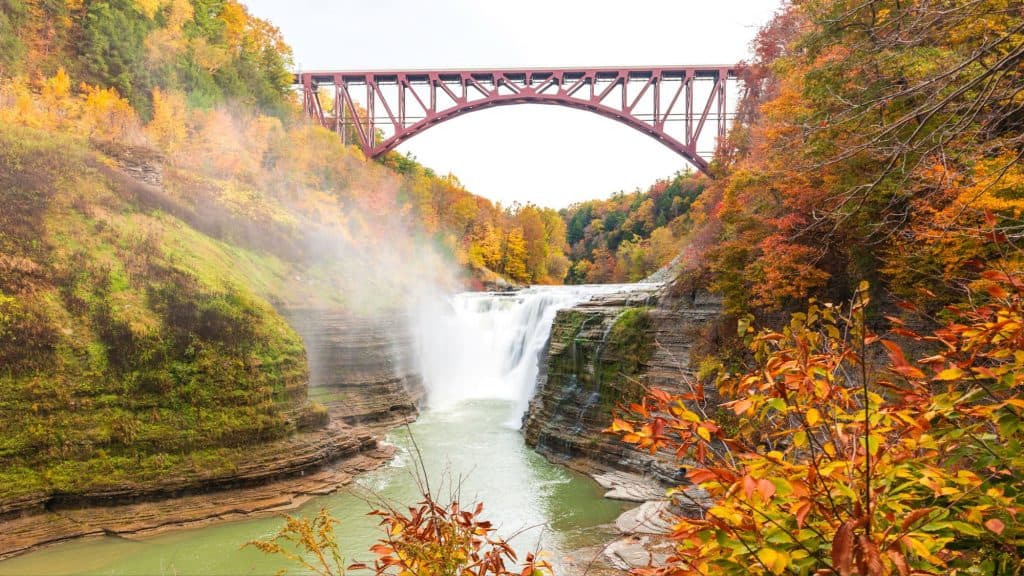 A powerful waterfall drops through a rocky gorge beneath a tall red bridge, with mist rising into the surrounding fall foliage.