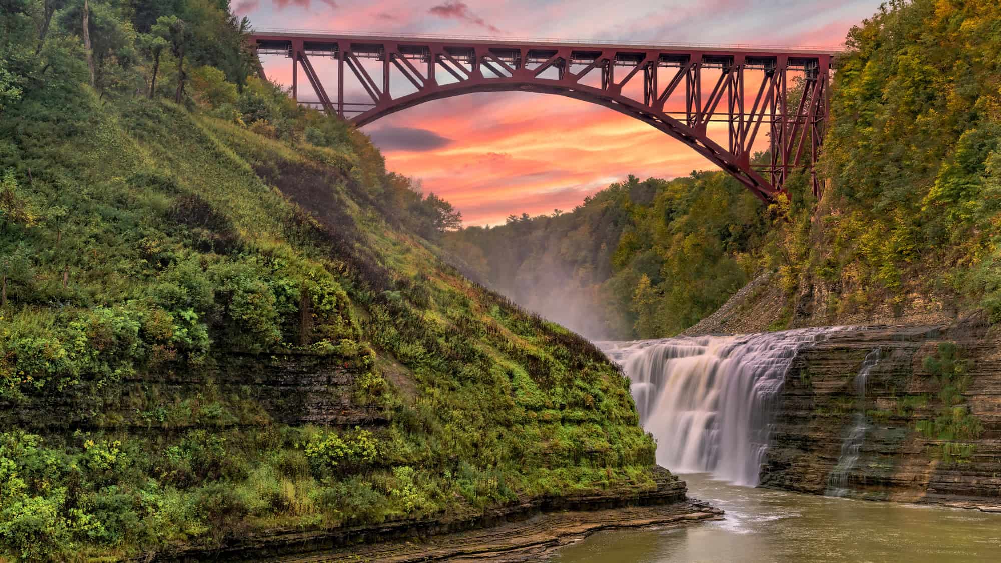 A dramatic red steel bridge arches over a powerful waterfall flowing through a gorge at sunset.
