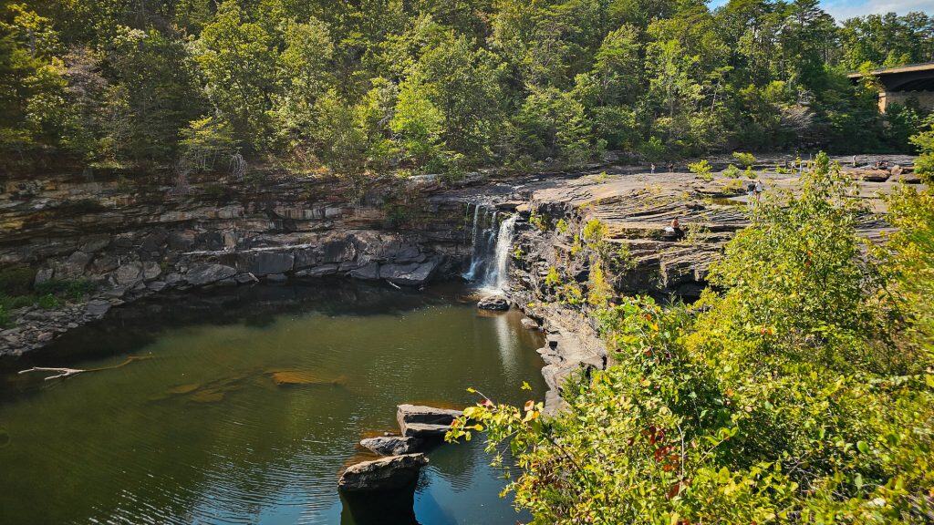 A small waterfall cascades into a calm pool surrounded by layered rock cliffs and lush greenery.