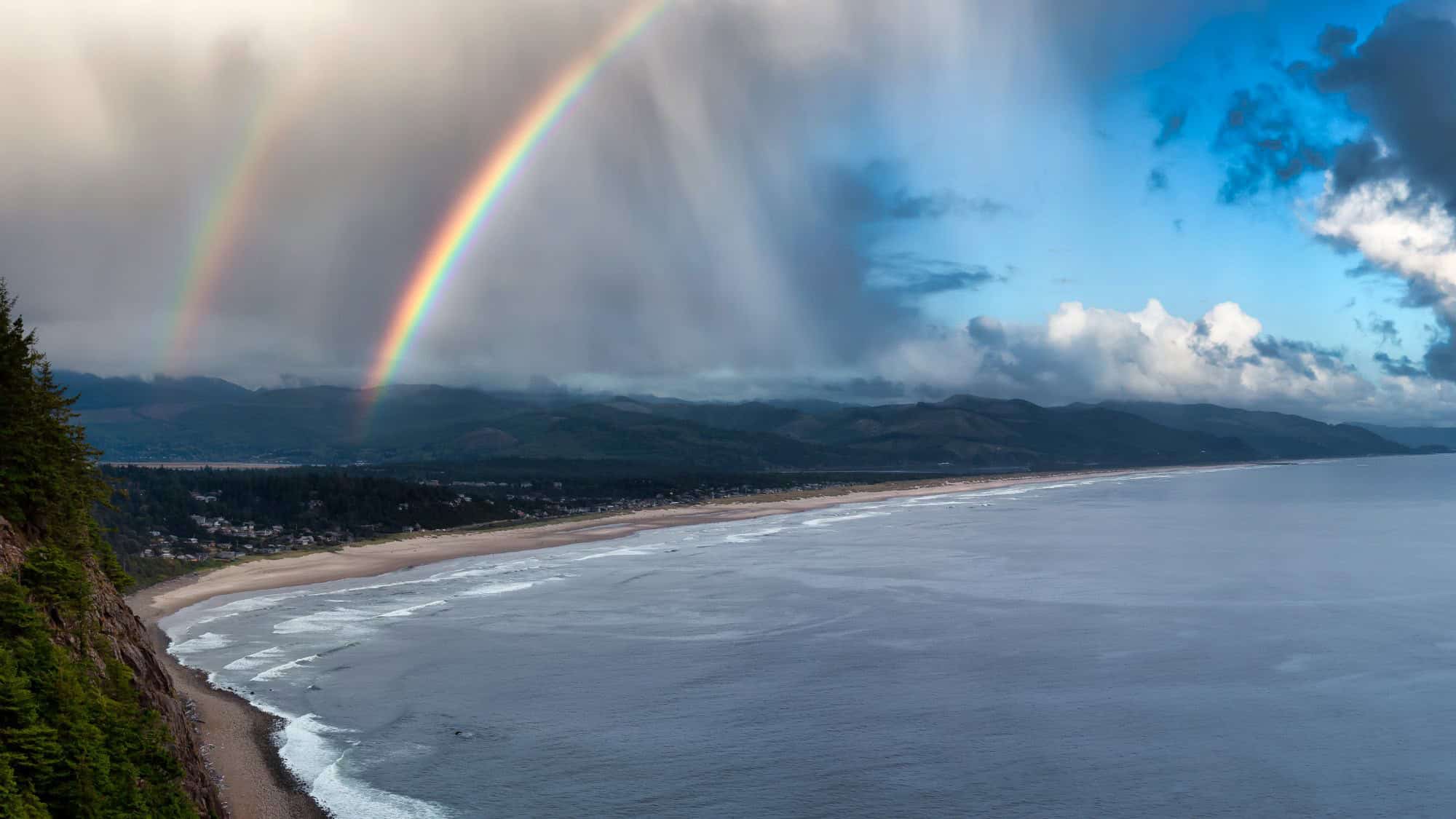 A vivid rainbow arches over the shoreline and mountains, with waves rolling onto the sandy beach under dramatic clouds.