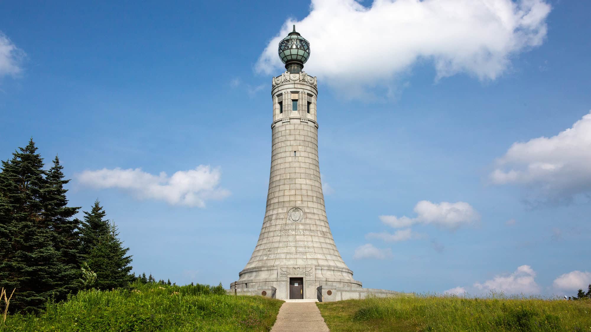A tall stone war memorial tower with a rounded base and glass beacon rises above a grassy hilltop under a bright blue sky.