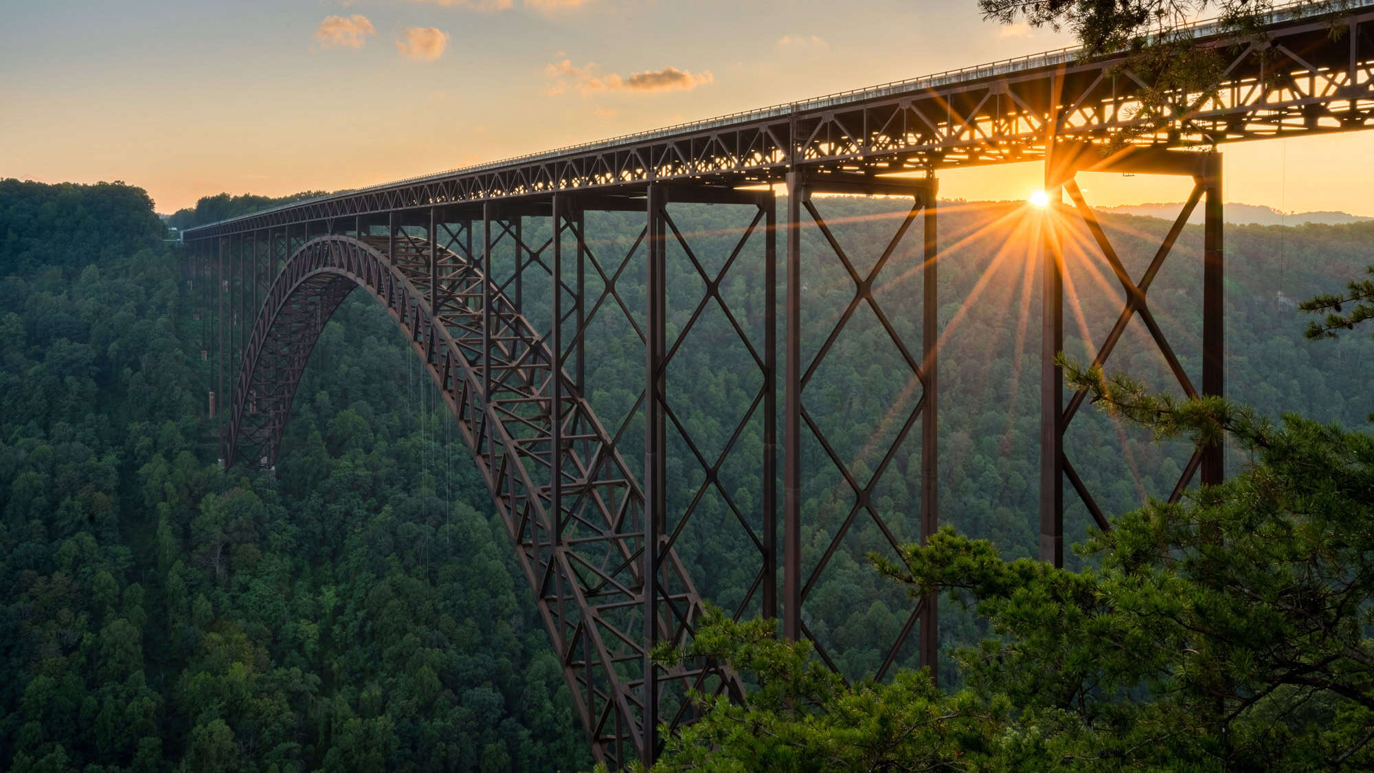The steel arch of the New River Gorge Bridge rises high above a forested canyon as the sun sets in a golden burst through its beams.