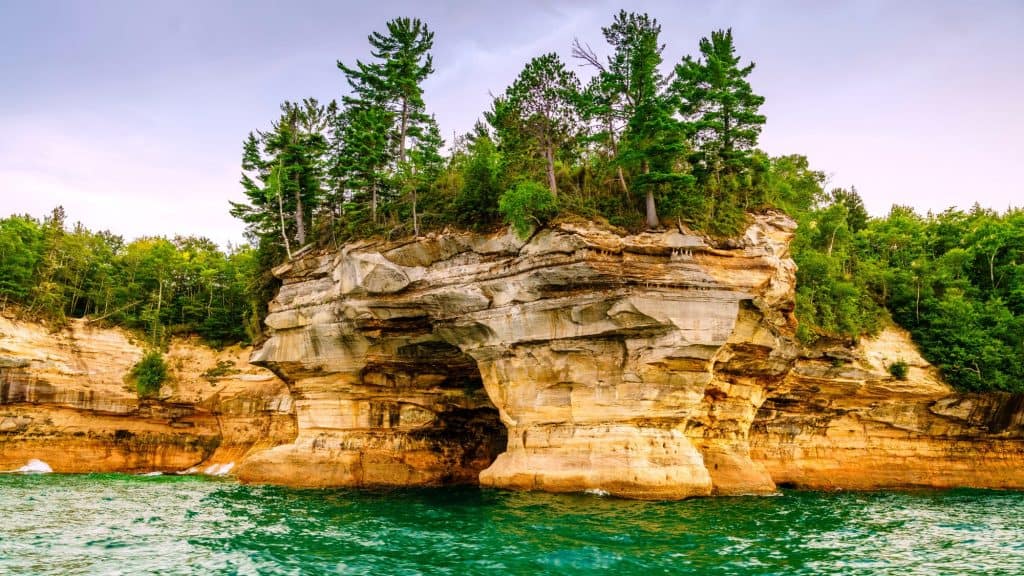 A striking sandstone cliff with layered hues of gold, orange, and beige rises above emerald waters, topped with dense green pines and forest.