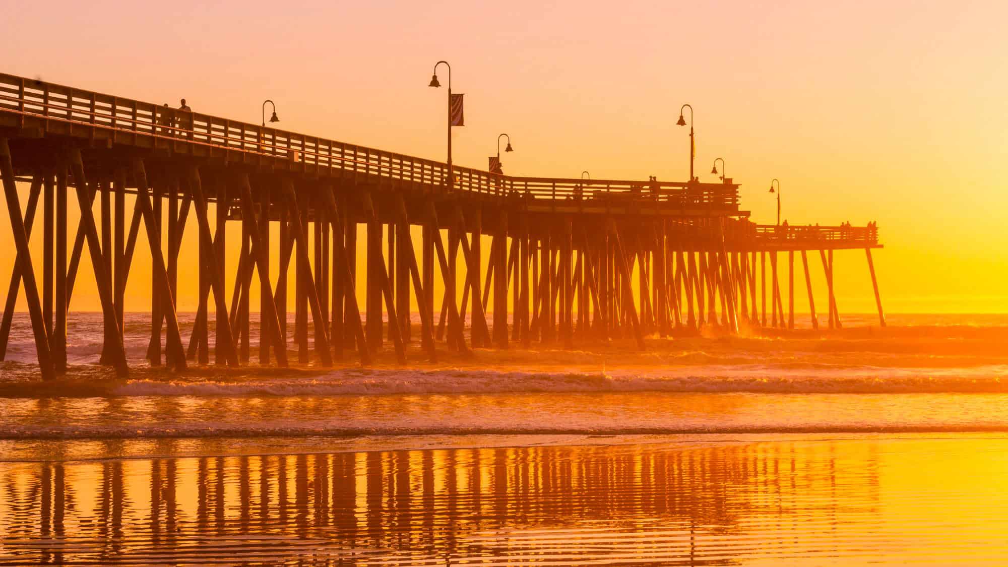 A wooden pier stretches into the ocean, glowing orange in the light of a brilliant sunset.