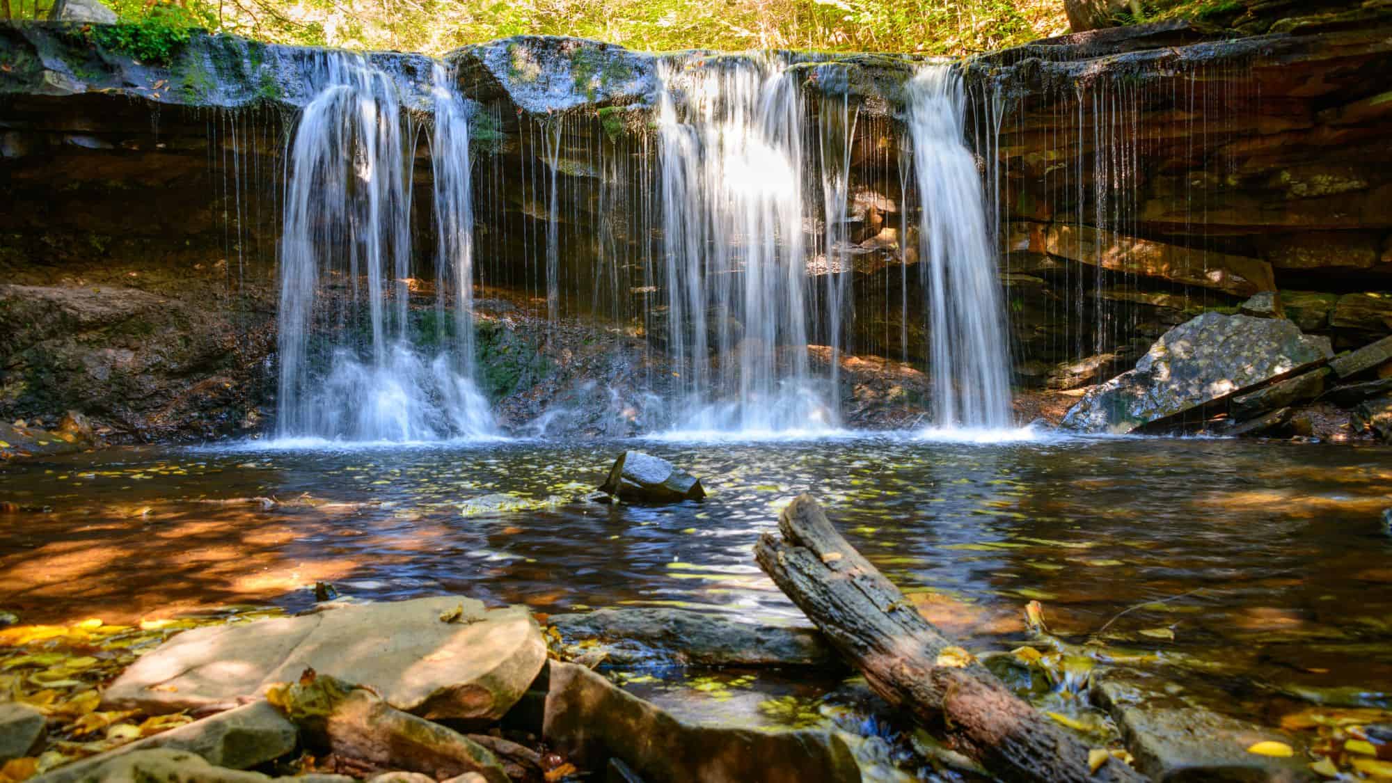 A towering waterfall plunges from a high rock cliff into a misty pool, framed by green trees and rugged canyon walls.