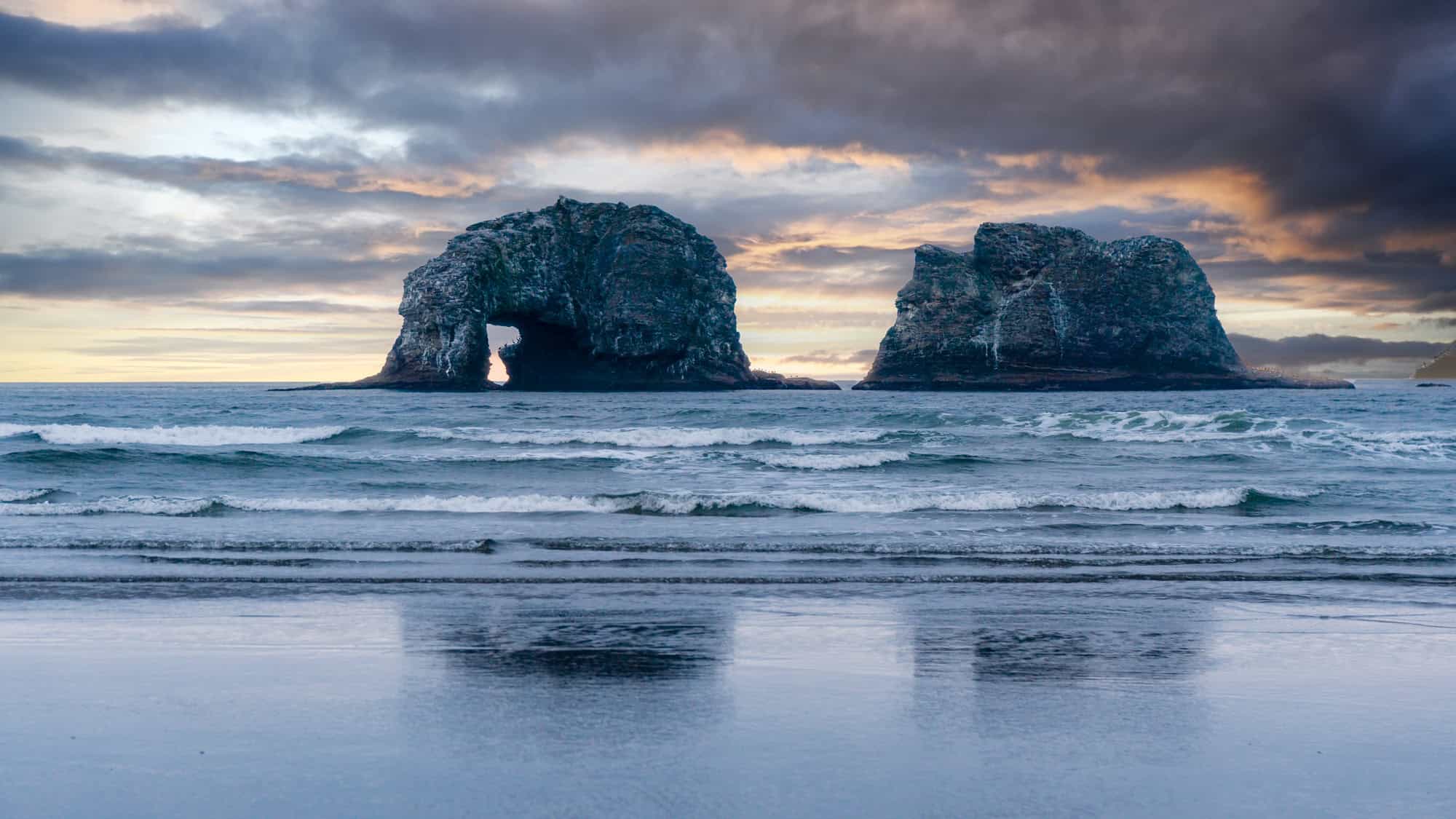 Ocean waves lap against the shore with two massive sea stacks rising from the water, one with a natural arch at its base.