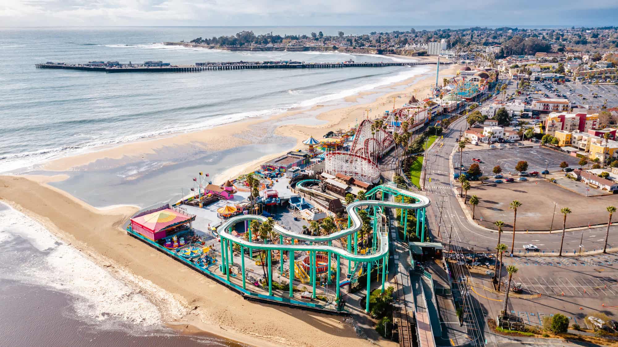 A colorful amusement park with roller coasters and rides lines the beach, with the pier stretching out into the ocean nearby.