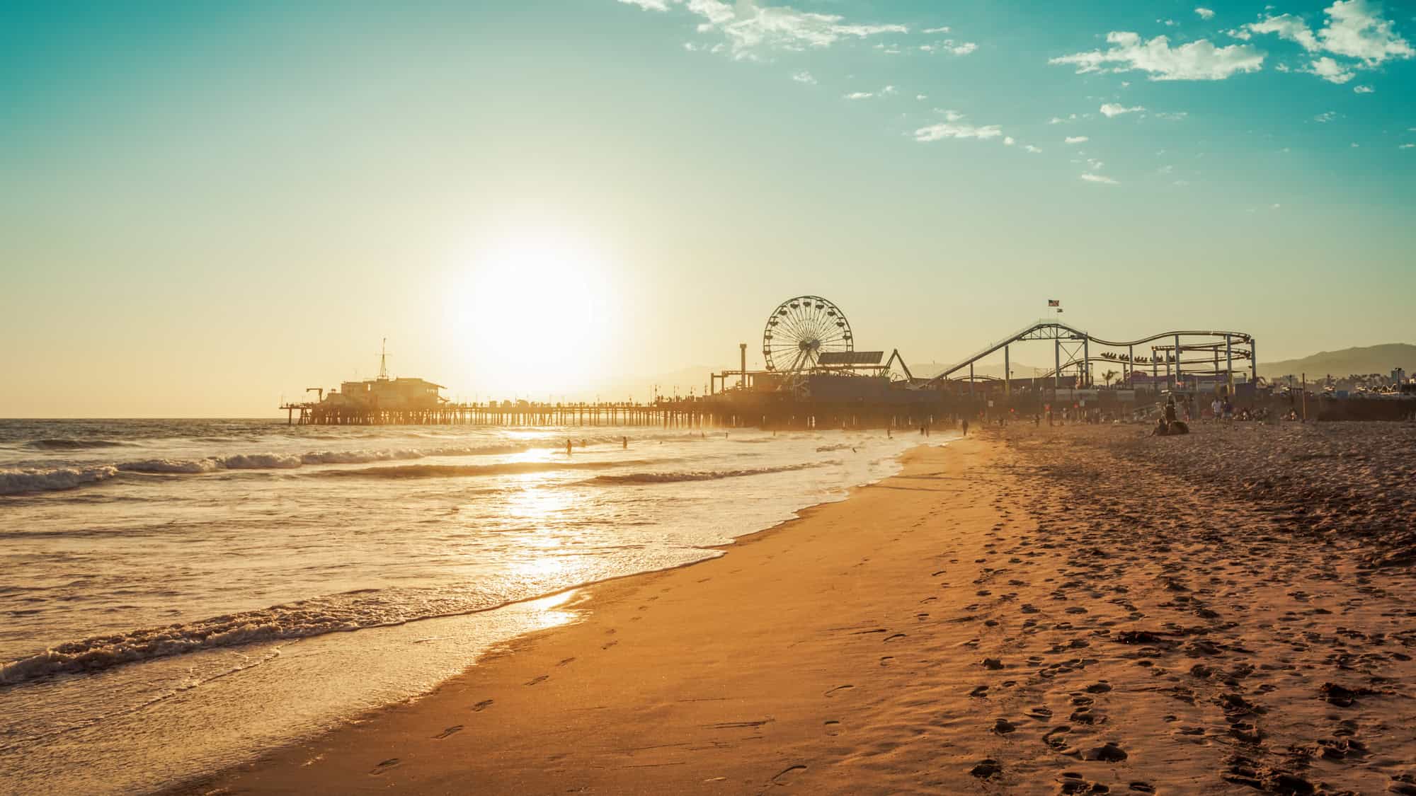 The sun sets over the ocean, casting golden light on the beach with the Santa Monica Pier’s Ferris wheel and roller coaster in the distance.