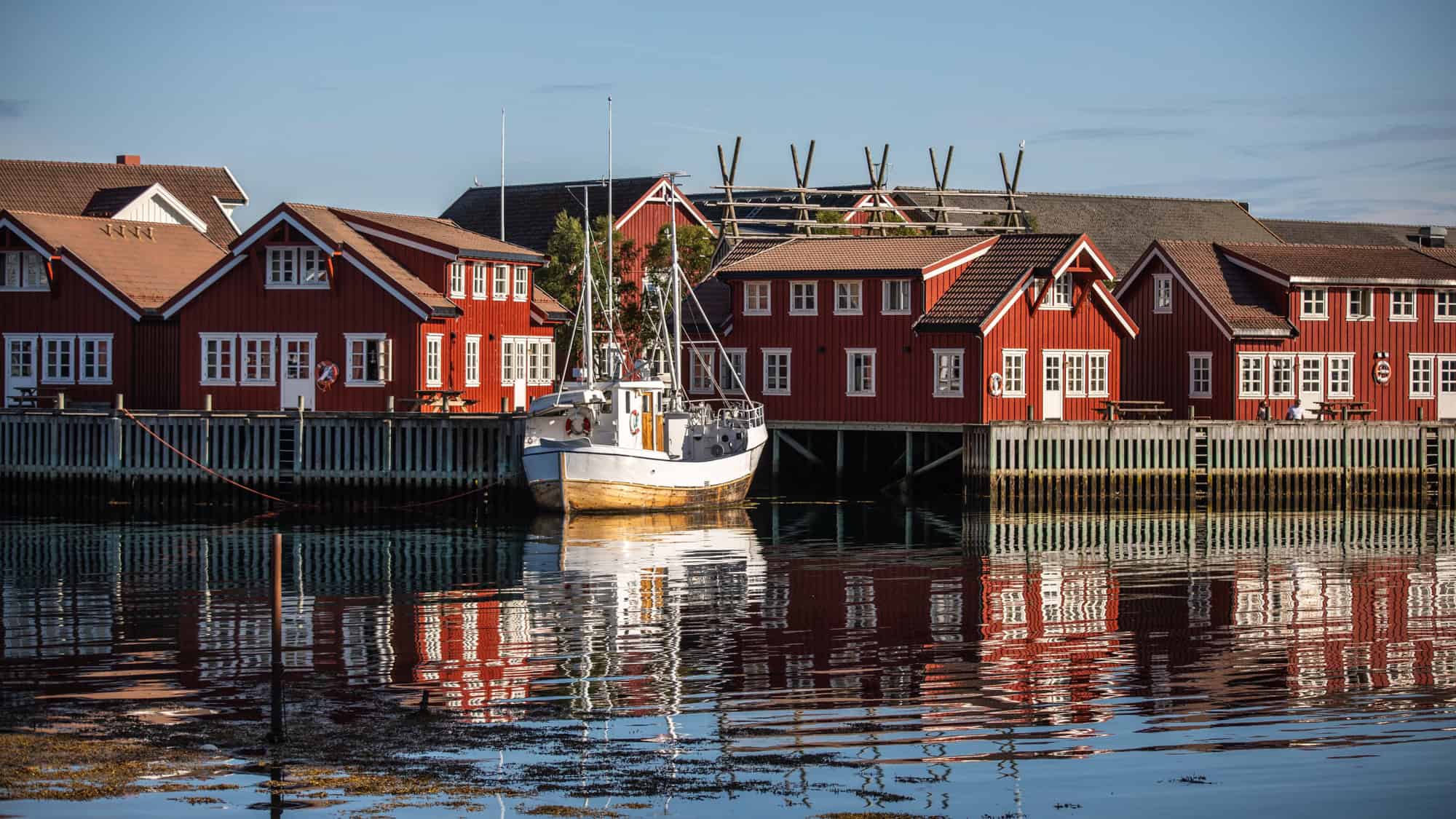 A cluster of red wooden waterfront houses reflects in the calm water, with a white fishing boat moored in front.