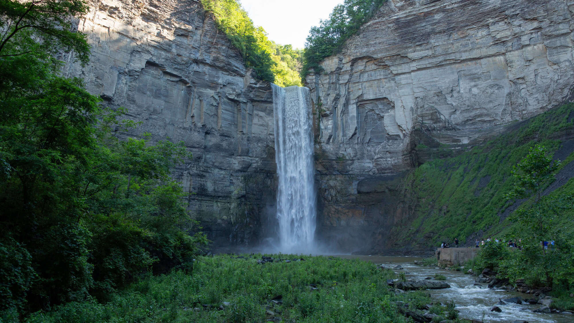 A towering waterfall plunges from a high rock cliff into a misty pool, framed by green trees and rugged canyon walls.