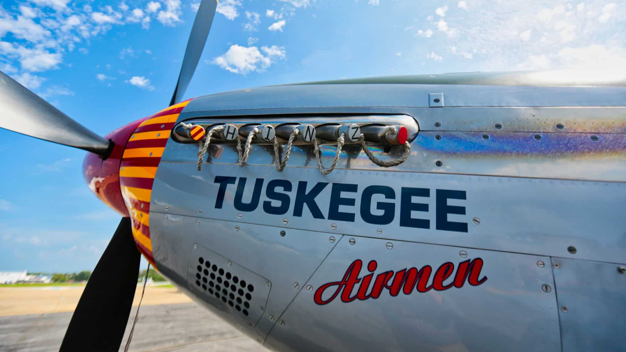 Close-up of a shiny silver aircraft with “Tuskegee Airmen” painted on the side, featuring a red and yellow striped nose and propeller.