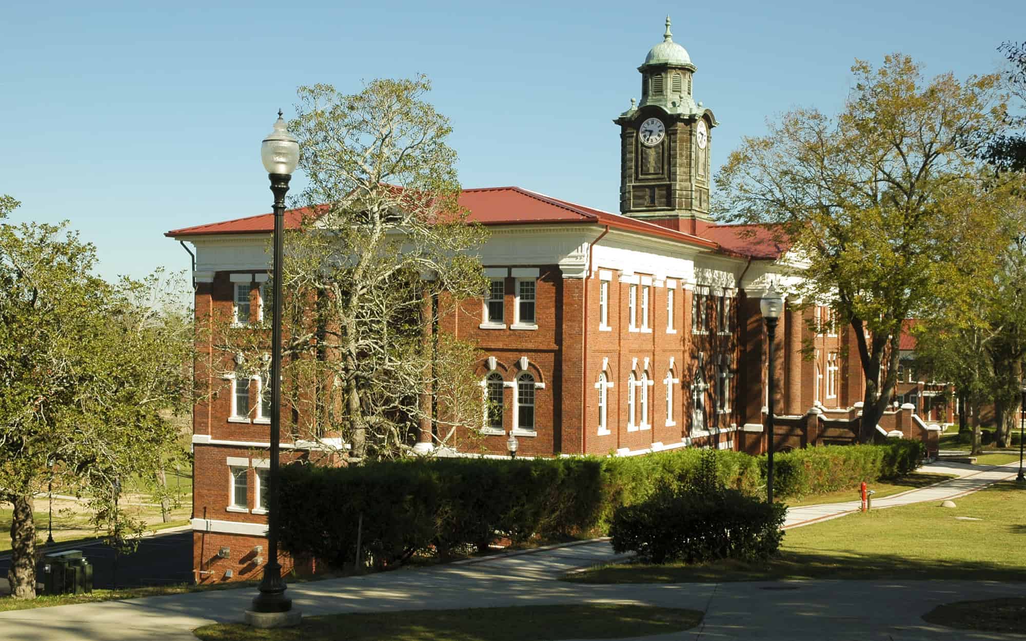 A stately red-brick building with a clock tower and red roof sits among trees on a sunny day.