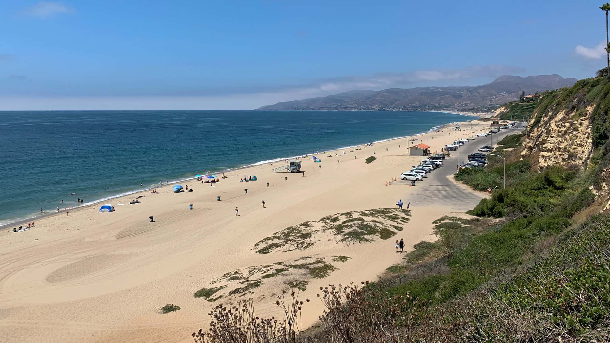 A wide sandy beach with scattered umbrellas and swimmers stretches along the ocean, backed by cliffs and rolling hills.