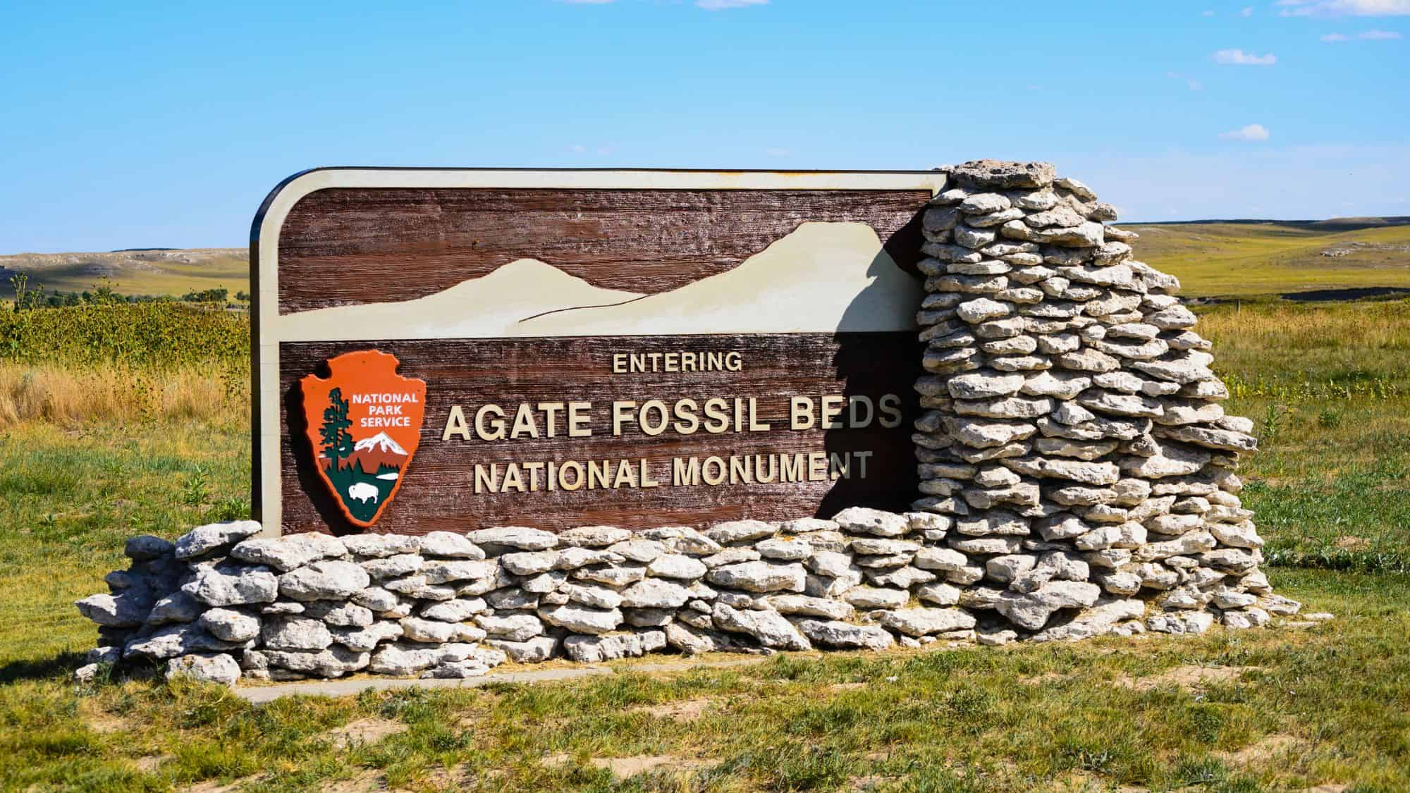 A National Park Service sign made of wood and stone marks the entrance to Agate Fossil Beds National Monument against a backdrop of grassy plains.