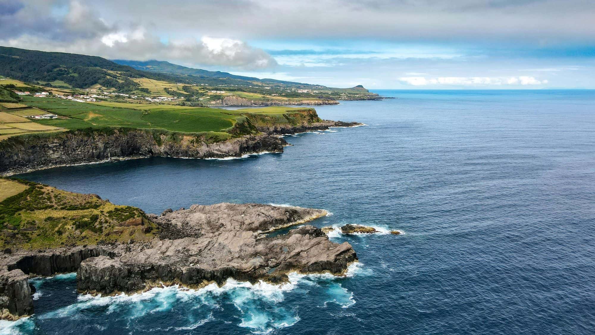 Crashing waves hit jagged volcanic rock formations along the coast, with green farmlands and rolling hills in the background.