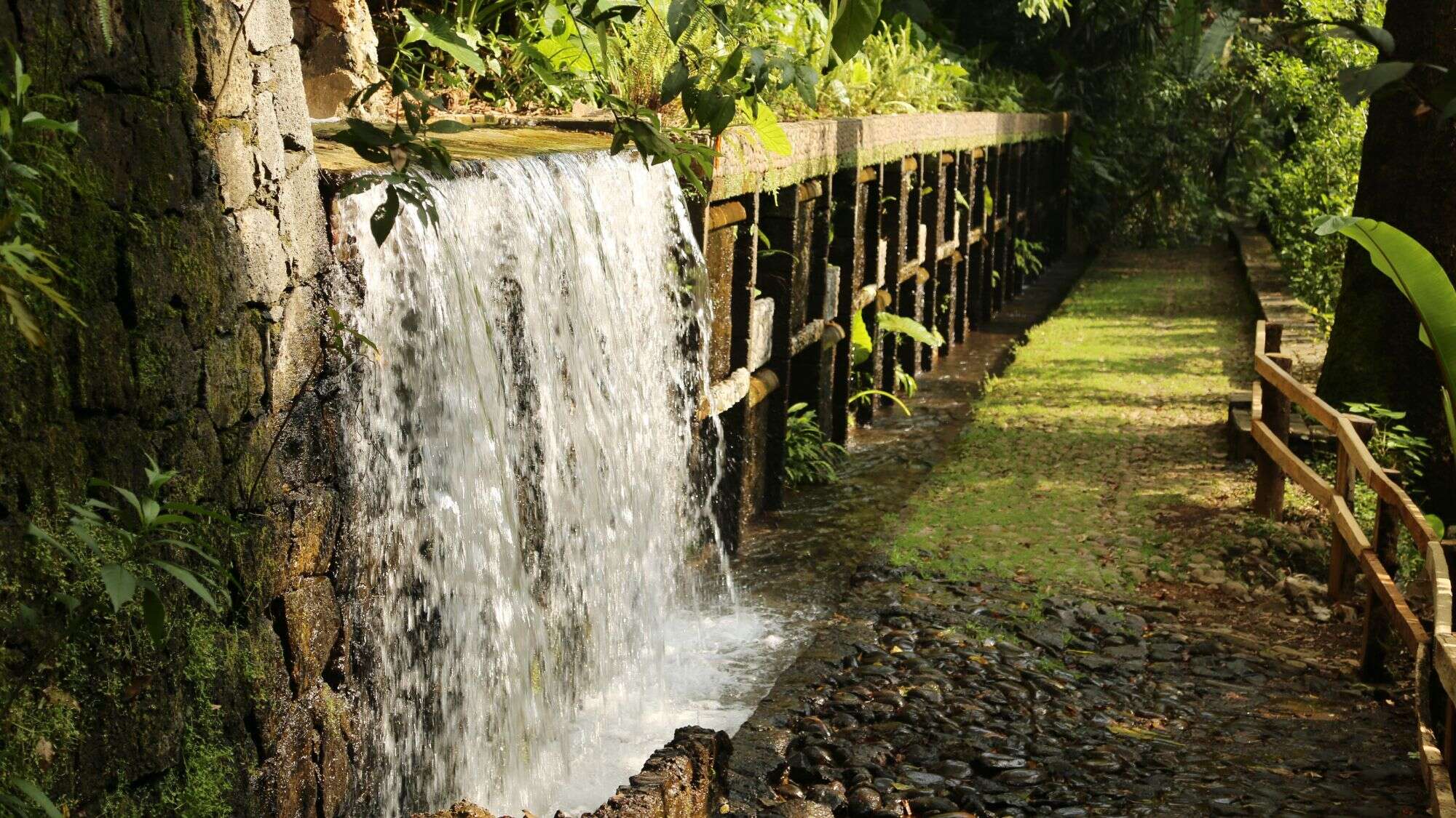 Water flows from a mossy stone aqueduct into a lush green forest path lined with a wooden railing.