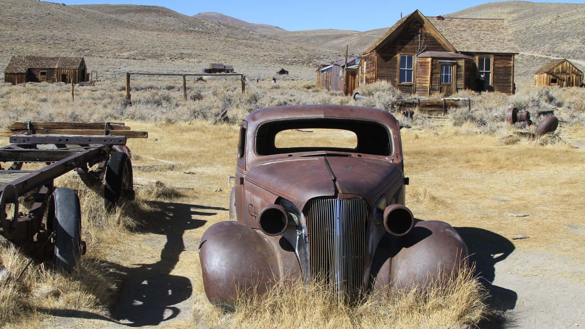 A decaying vintage car sits in dry grass near old wooden cabins in Bodie, California, capturing the town’s eerie frozen-in-time atmosphere.