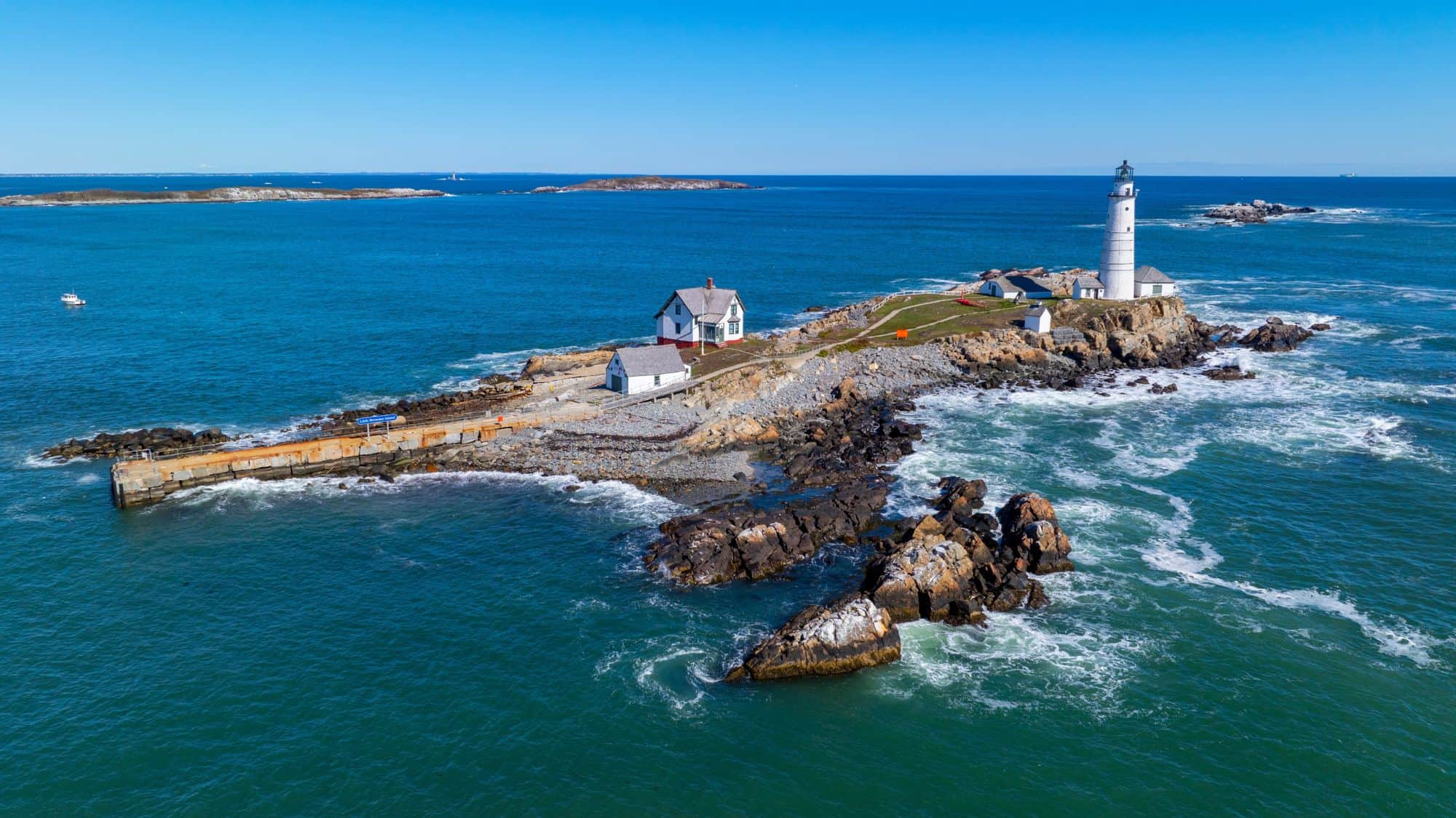 A tall lighthouse stands on a rocky island surrounded by ocean, with several buildings and a pier dotting the rugged shoreline.