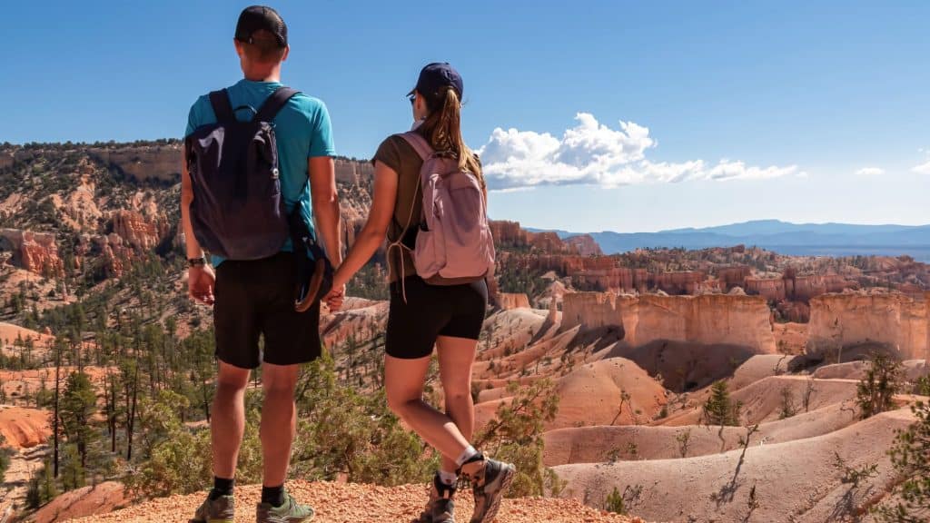 A man and woman wearing backpacks hold hands while admiring the red rock formations of a canyon under a bright blue sky.