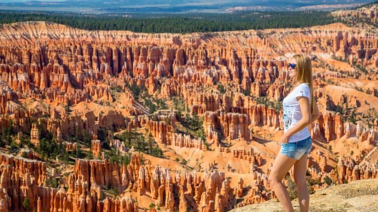 A woman in sunglasses and shorts gazes out over the dramatic red rock formations of Bryce Canyon under a clear blue sky.