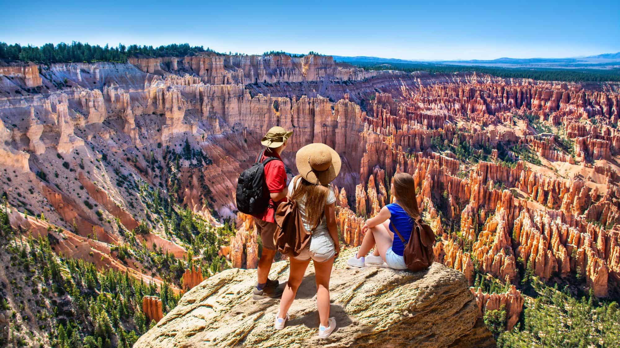 Three hikers stand on a cliff edge admiring the vast amphitheater of orange rock spires and hoodoos at Bryce Canyon.