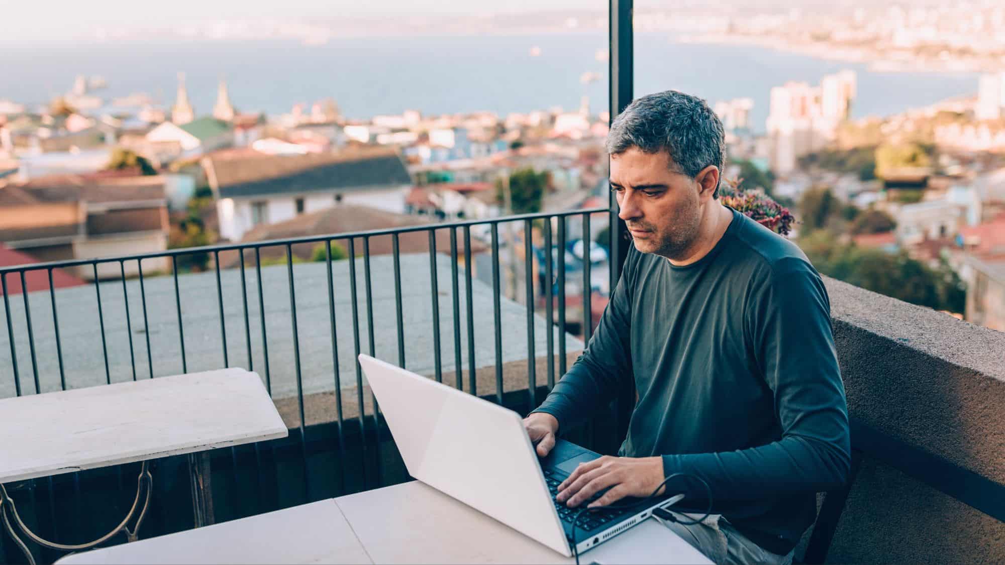 A man works on a laptop at an outdoor table overlooking a coastal city with the ocean in the distance.
