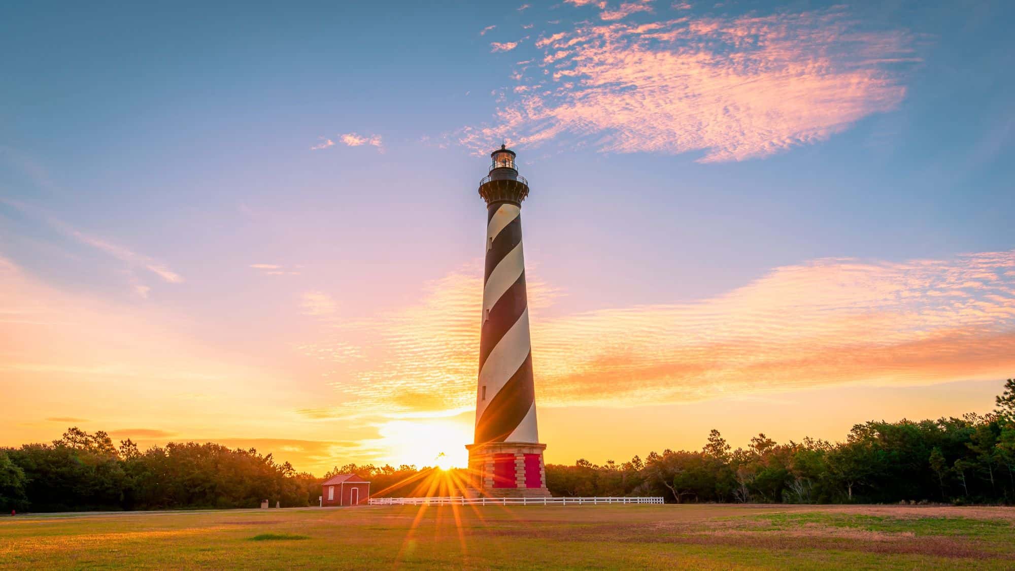 A tall black-and-white spiral-striped lighthouse rises dramatically against a colorful sunset sky, with sunbeams flaring from the horizon.