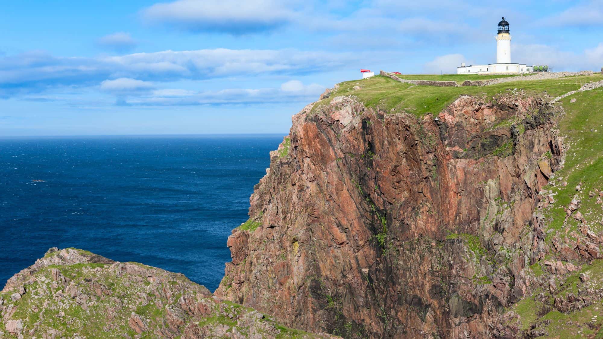 A white lighthouse stands atop steep red sea cliffs, surrounded by green grass and overlooking the deep blue ocean.