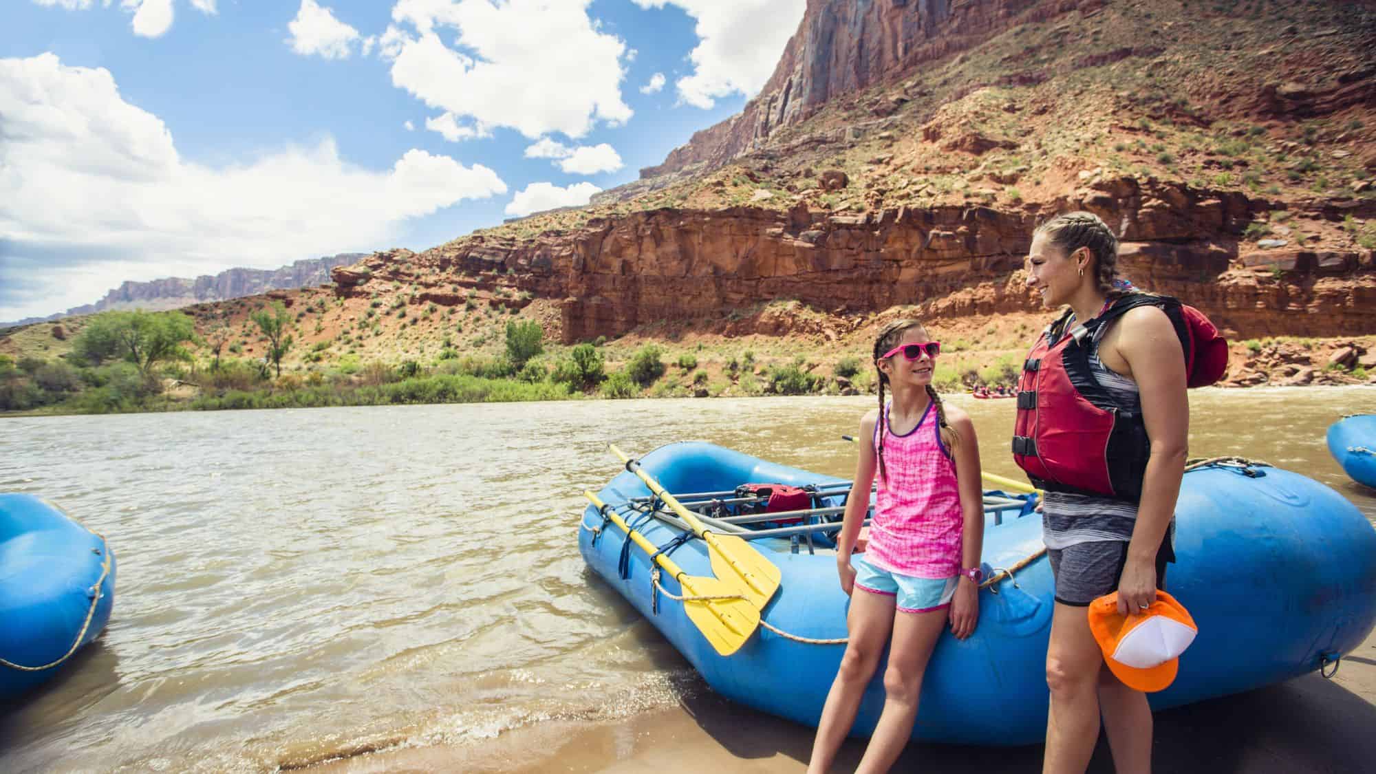 A woman and child in life vests stand beside a blue raft on a calm river with red rock cliffs in the background.