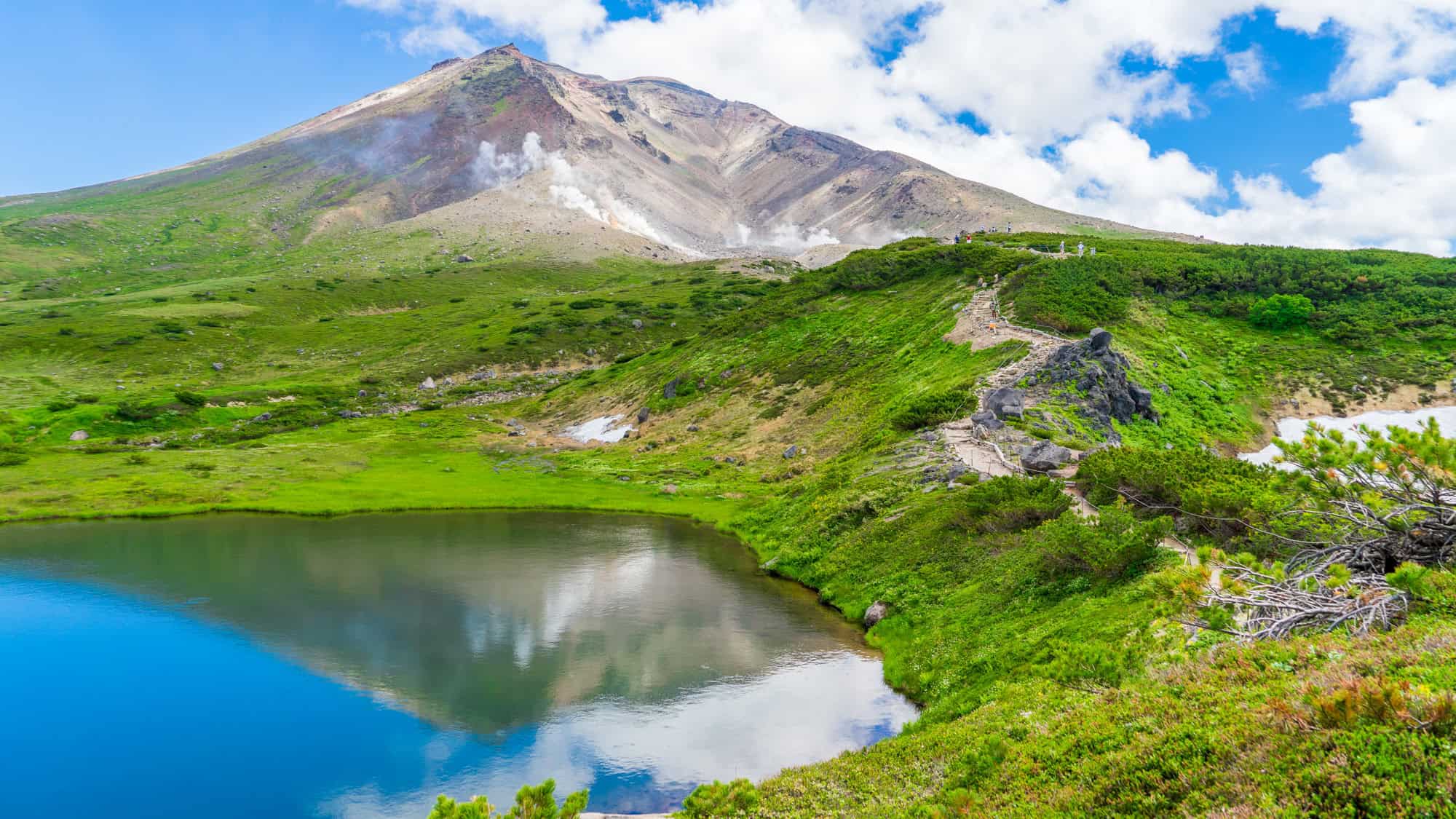 A steaming volcanic slope looms above a bright green valley and reflective blue lake.