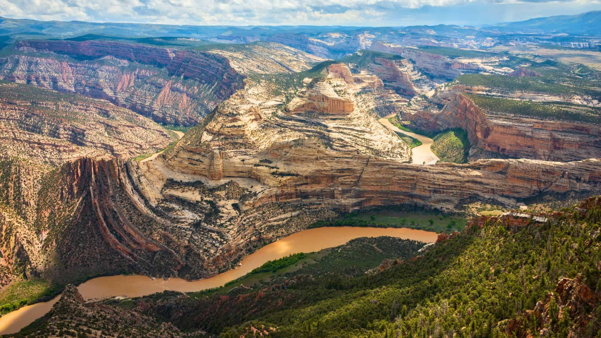 A winding river cuts through layered red and tan canyon walls, creating dramatic curves and cliffs in a vast desert landscape.