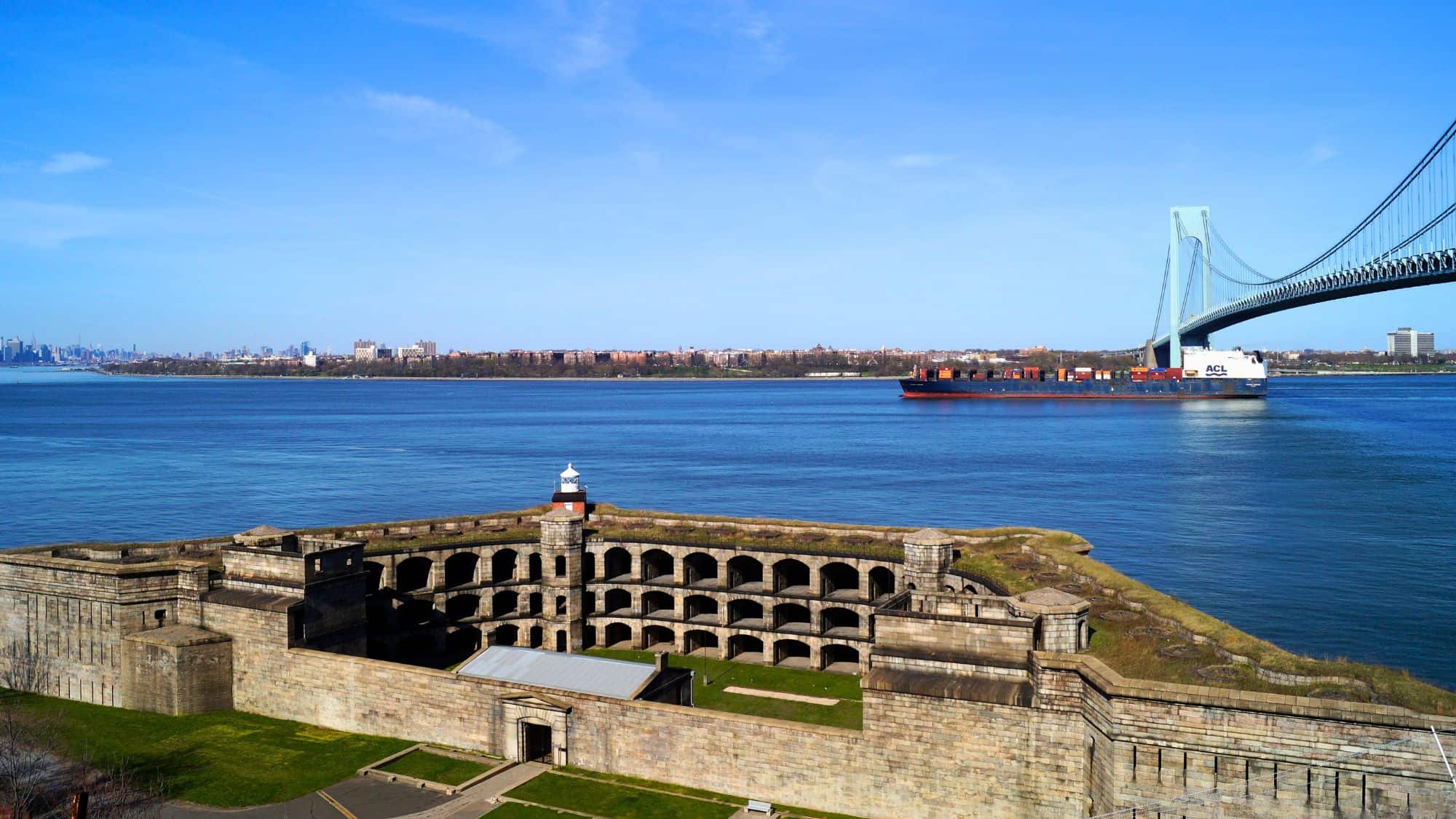 A historic stone fort with a small lighthouse is seen beneath the Verrazzano-Narrows Bridge, with a cargo ship sailing past in the harbor.